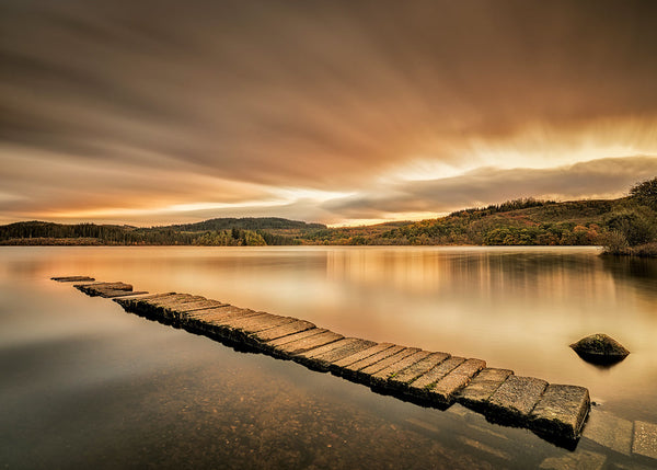 A long-exposure landscape photograph of a stone stepping stone path leading out of a calm lake toward distant hills under a dramatic golden sky.
