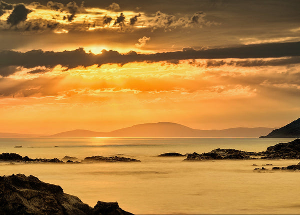 Ballycastle sunrise over calm sea, County Antrim Northern Ireland, golden light reflecting on water with distant coastline and rocky foreground
