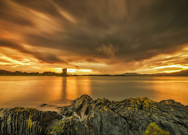 A dramatic landscape photograph of a rocky shoreline at sunset, with golden-orange clouds streaking across the sky, calm water reflecting the light, and a distant castle on the horizon.