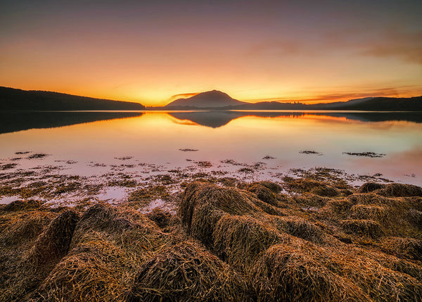 Connemara lake at sunset with still water reflecting mountain silhouette and warm sky by Irish landscape photographer John Taggart