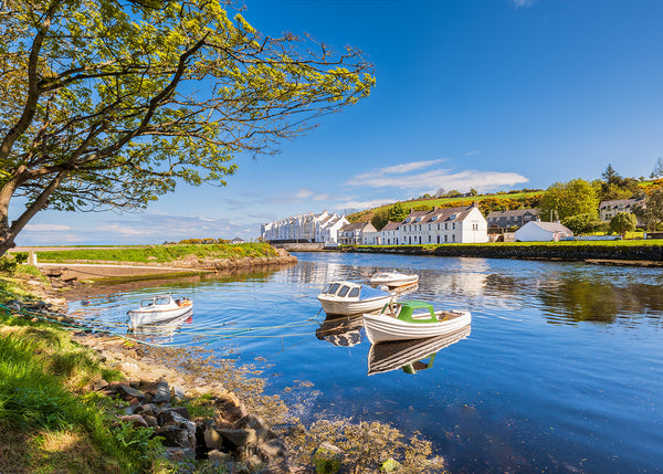 A peaceful view across Cushendun Harbour by John Taggart Landscapes on the beautiful Causeway Coast of County Antrim, Northern Ireland. Small boats rest on the calm, reflective water while the village’s traditional whitewashed buildings glow beneath a clear blue sky. Framed by the sweeping branches of a mature tree, the scene captures the quiet coastal charm for which Cushendun is loved.