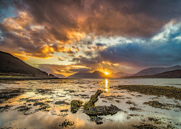 A dramatic sunset over a calm loch, featuring fiery orange light breaking through heavy dark clouds, a bright sunburst setting behind distant mountain peaks, and seaweed-covered rocks resting in the shallow, reflective water of the foreground.