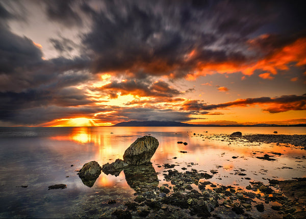Irish coastal sunset with dramatic clouds, glowing reflections on calm sea and rocks in foreground