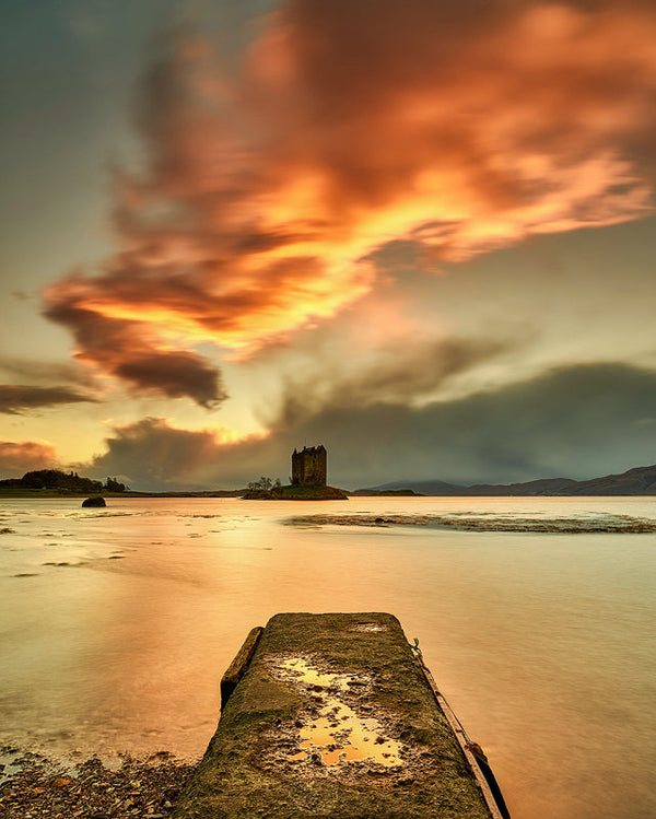 A dramatic fiery orange sunset sky over a calm body of water, featuring a weathered stone pier in the foreground leading the eye toward a solitary, square castle on a small island, with faint mountains in the background.