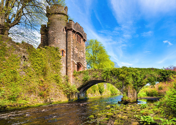 A historic stone tower and an ivy-covered arched bridge spanning a shallow, rocky river, surrounded by vibrant green trees under a bright blue sky.