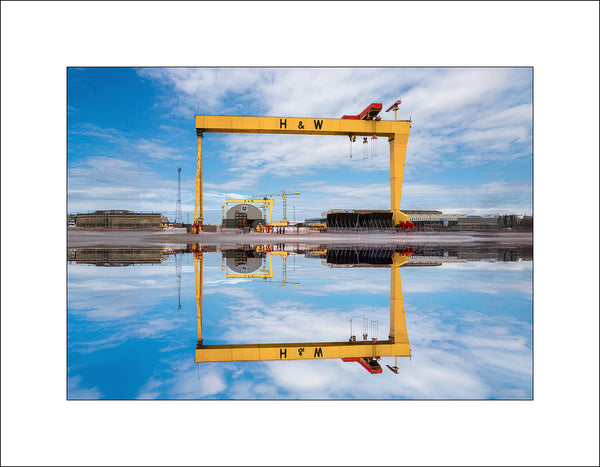 Standing tall against a vivid Belfast sky, the iconic Harland & Wolff cranes—Samson and Goliath—are perfectly mirrored in water, creating a striking symmetry. A powerful symbol of the city's shipbuilding heritage, this image captures the strength, scale, and beauty of Belfast’s industrial past and present. A moment of calm in the heart of the docks. By John Taggart Landscapes