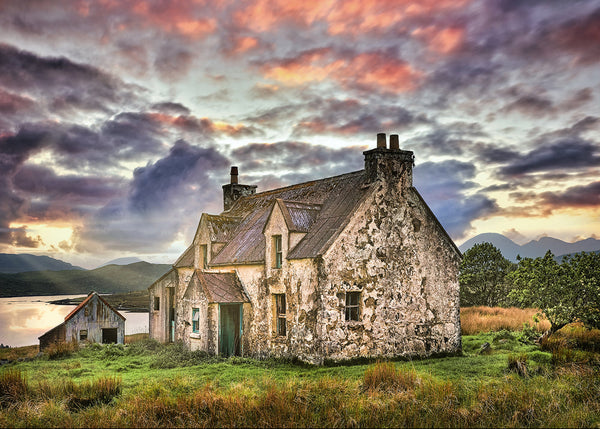 A weathered, historic stone cottage with peeling white paint sits in a grassy green field near a calm loch, set against a dramatic sky filled with vivid pink, orange, and dark purple clouds.