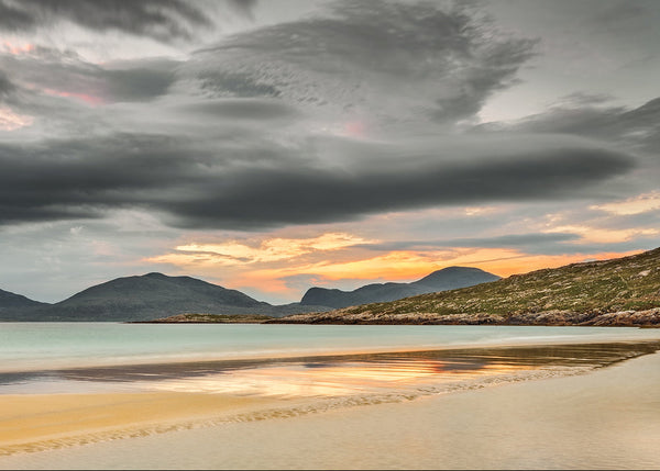 A dramatic coastal sunset featuring smooth, wet sand reflecting warm golden light, calm turquoise water, and dark mountain silhouettes beneath massive, swirling gray clouds.