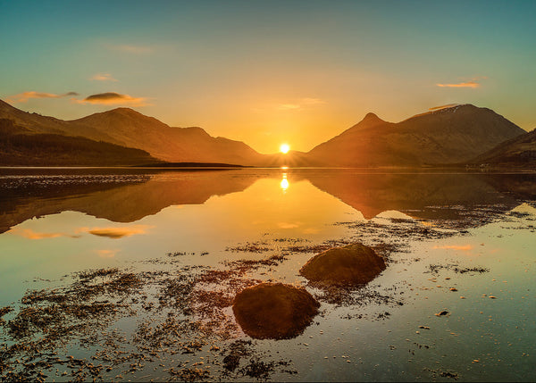 A radiant golden sunset over a calm loch, featuring a bright sunburst setting between distant mountain peaks, perfectly mirrored in the still water, with two prominent dark rocks in the shallow foreground.