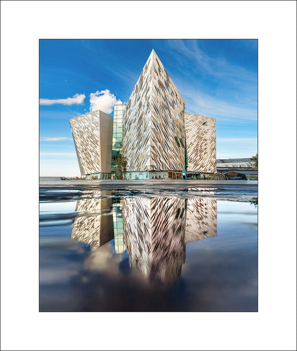 Striking architectural photography of Titanic Belfast, captured with perfect reflection and crisp detail under a blue sky. This iconic image from Northern Ireland showcases the modern design and cultural significance of Belfast's Titanic Quarter. A fine art print by John Taggart Landscapes, ideal for home or office decor.