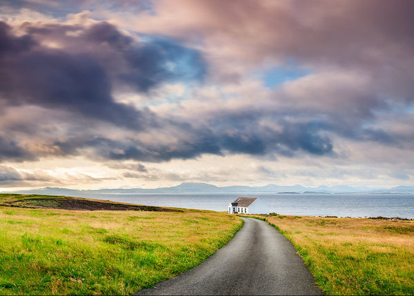 Winding road leading to a small building with a dramatic sky over water and mountains.